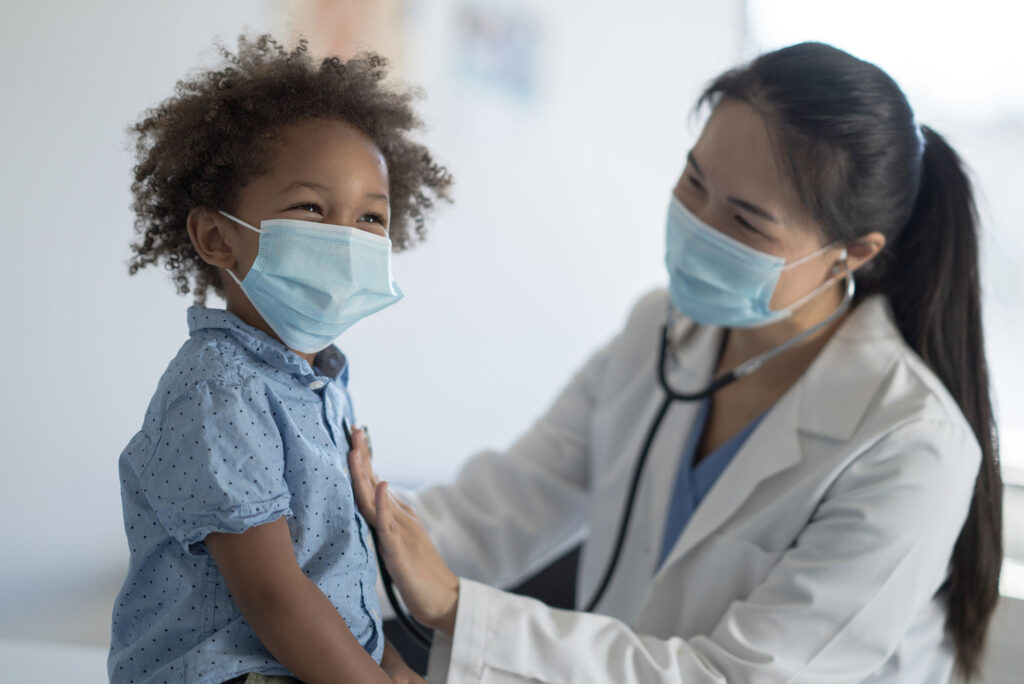 A female doctor of Asian decent holds her stethoscope to a little boys chest as she listens to his heart. The little boy is dressed casually and smiling as he sits still for the appointment. Both the patient and the doctor are wearing medical masks to protect them from COVID.